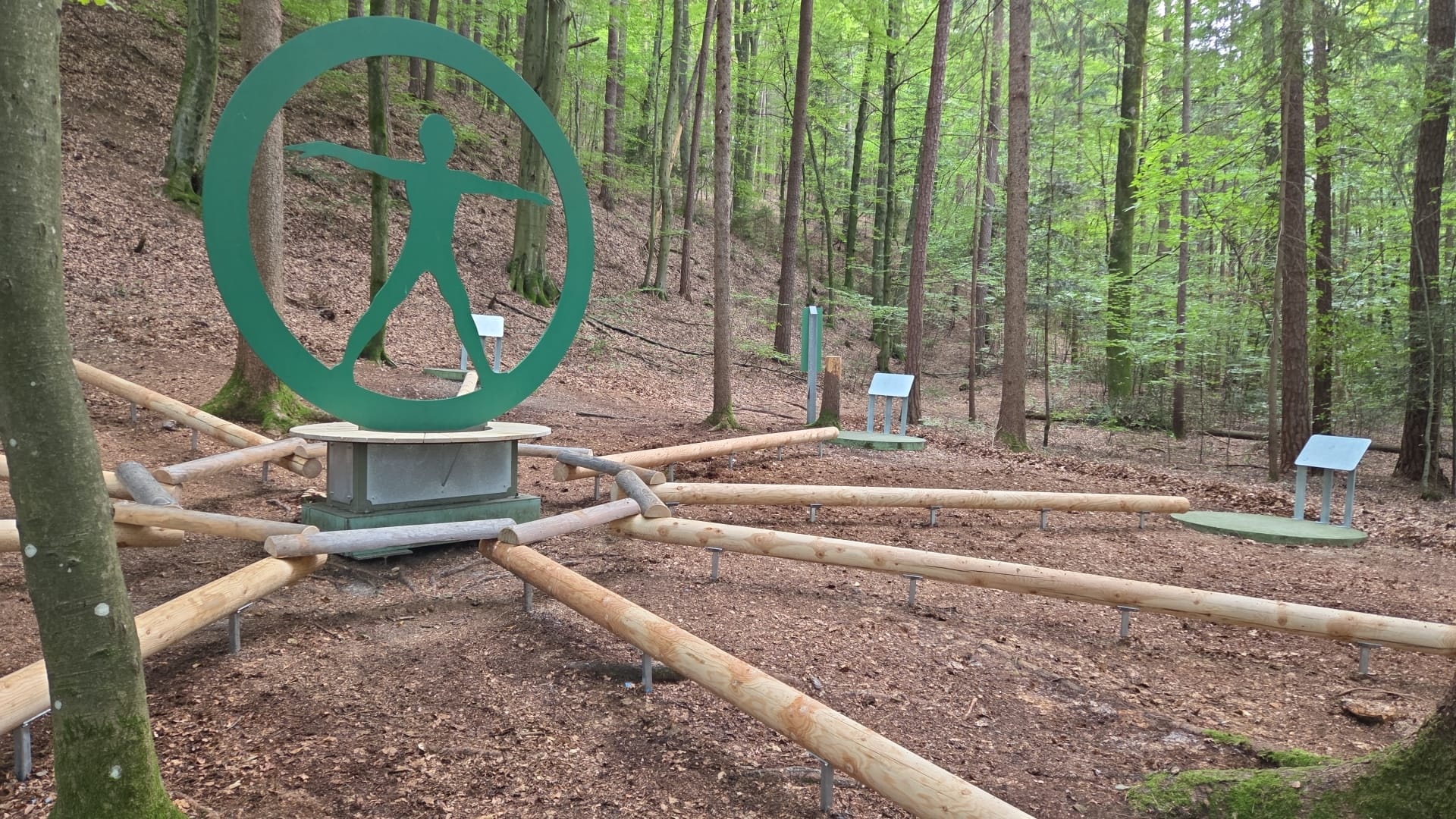 Station 8 - Sonnengeflecht Das Bild zeigt die Erlebnisstation 8: Eine grüne Skulptur bildet den Mittelpunkt von dem 9 runde Hölzer zu Tafeln mit Entspannungsübungen zeigen.